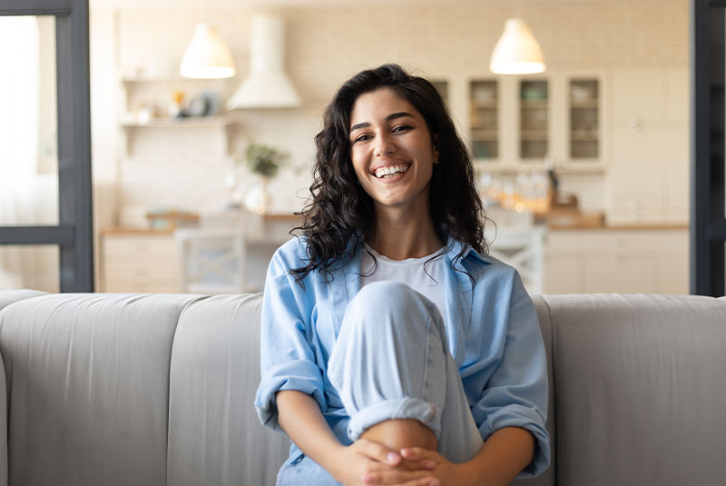 Portrait of pretty young brunette woman smiling and looking at camera, sitting on couch at home